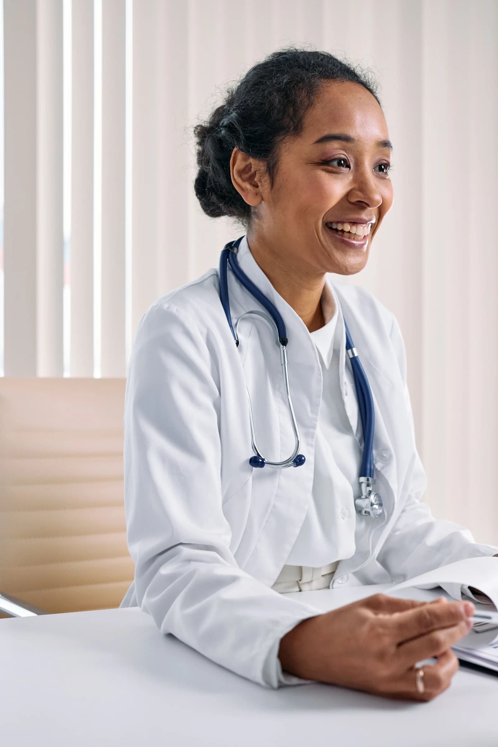 A Black female doctor in a white coat and stethoscope at her clinic desk.